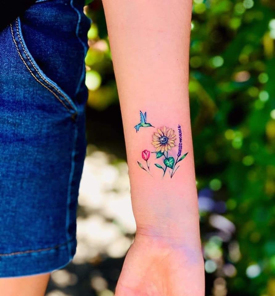 39 Tatuagem de beija-flor no antebraço mordendo girassol e flor de tulipa lavanda