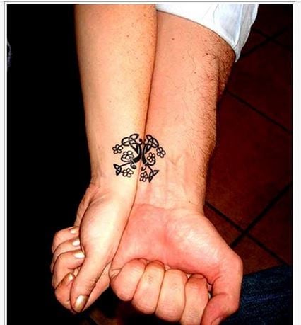 Small and complementary tattoos for couples on wrists, curled ornaments with little black flowers in the shape of a triqueta