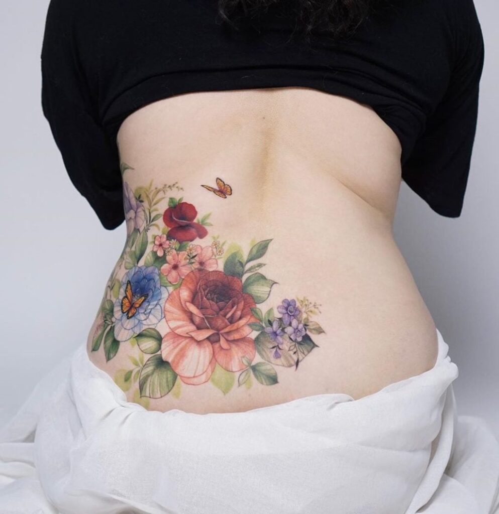 Tatuajes en espalda baja parte izquierda gran arreglo floral que sube por el costado del vientre rosas rojas flores azules mariposas hojas flores rosas