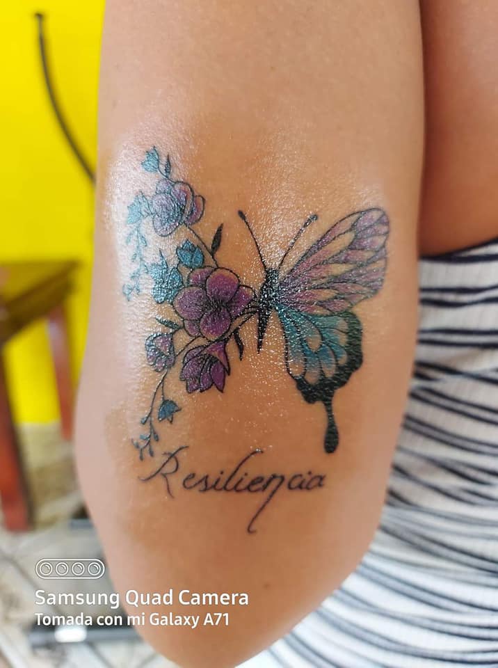 Les tatouages les plus aimés des femmes beau papillon dans les tons violet et bleu clair avec le mot résilience et petites fleurs