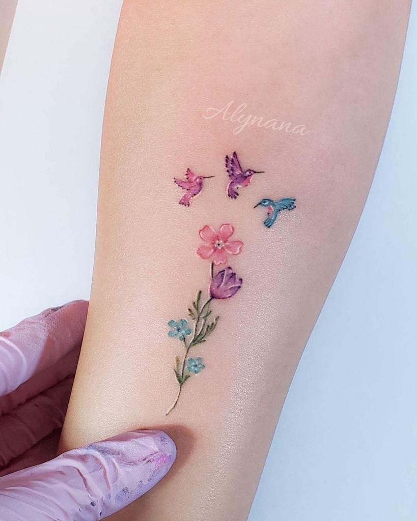 12 Estudio Alynana Tattoo CDMX Drei Kolibris, eine rosa-violette und blaue Blume, die drei Kinder auf dem Unterarm darstellen