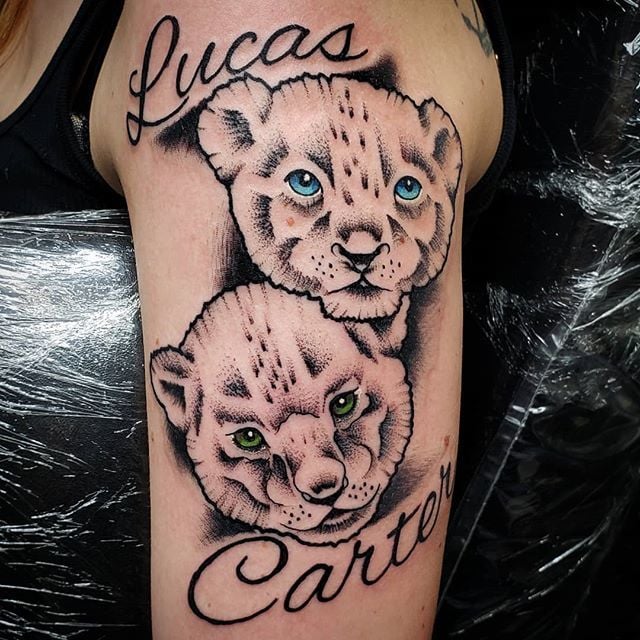 Tattoo von Leona und ihren Jungen. Gesicht von zwei Welpenkindern, eines mit grünen Augen, das andere mit hellblauen Augen und den Namen Lucas und Carter