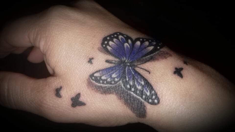 Tattoos für Frauen am beliebtesten: Violett-blauer Schmetterling auf der Hand mit kleinen schwarzen fliegenden Schmetterlingen