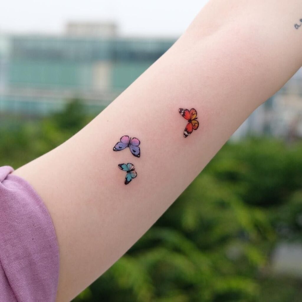 Tatuagens de pequenas borboletas laranja violeta azul claro no antebraço