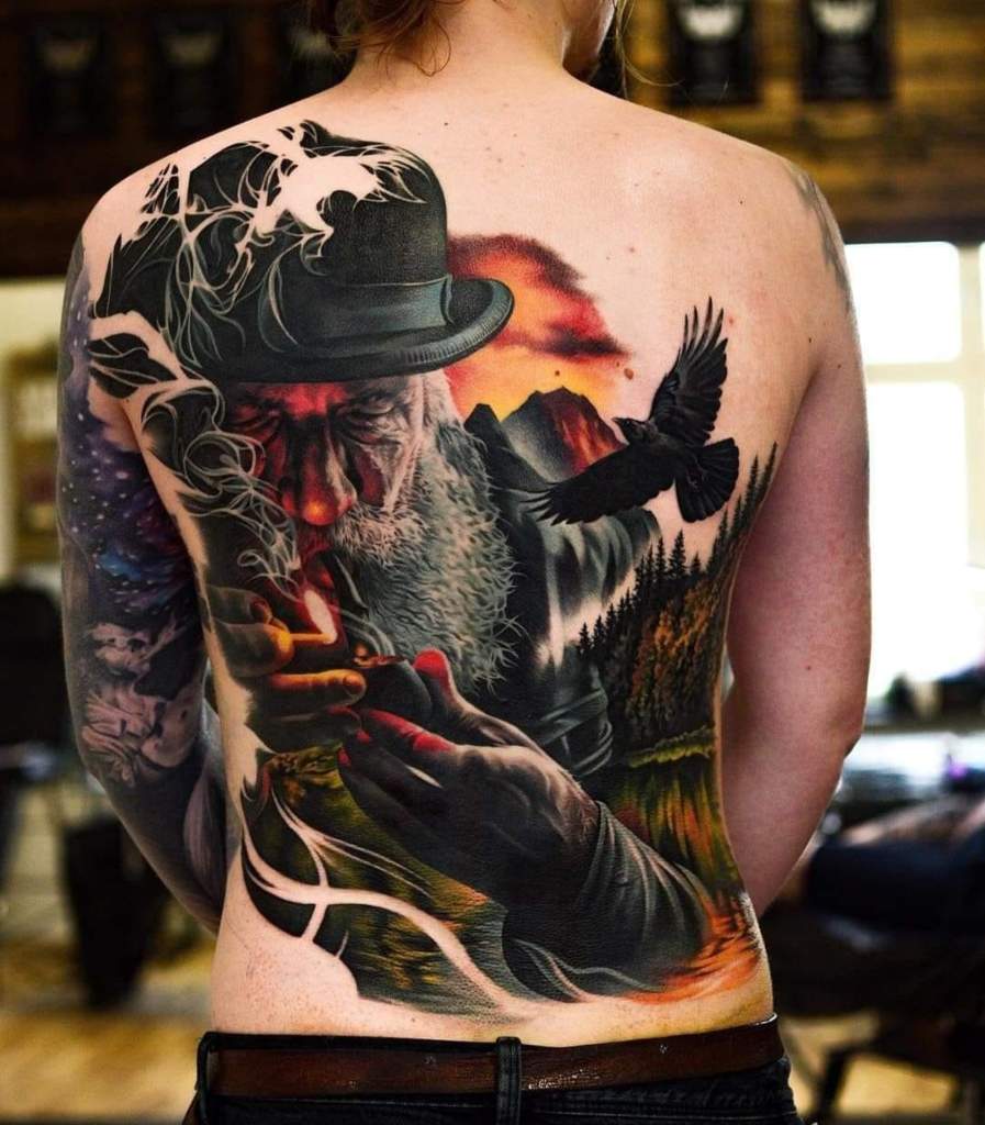 4 TOP 4 Homme tatoué réaliste avec barbe et chapeau fumant une pipe avec allumette allumée derrière un paysage de montagnes avec aigle sur le dos