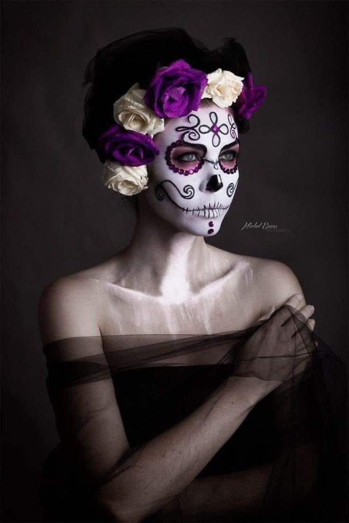 33 La Catrina-Kostüme, Make-up mit Cempasuchil-Blumensymbolen im Gesicht, Details in Lila, Stirnband mit lila und weißen Blumen, tief ausgeschnittenes schwarzes Kleid