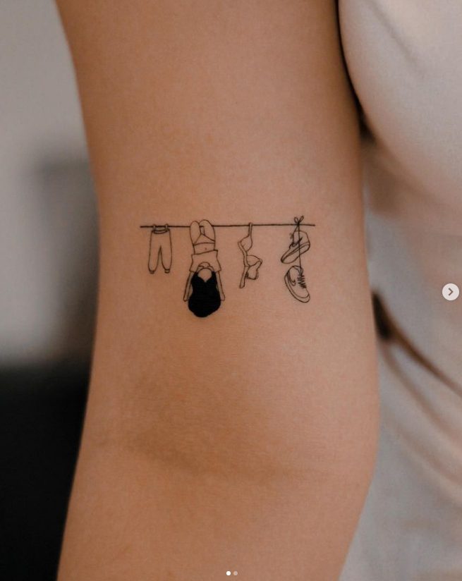 47 tatuaggio dell'artista Nhi.ink di una ragazza che gioca su uno stendibiancheria con scarpe da ginnastica e vestiti sul gomito