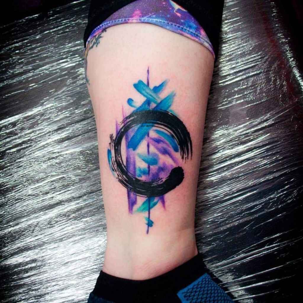 Tatuaje Circulo Zen trazos de acuarela de fondo celestes y violetas en pantorrilla