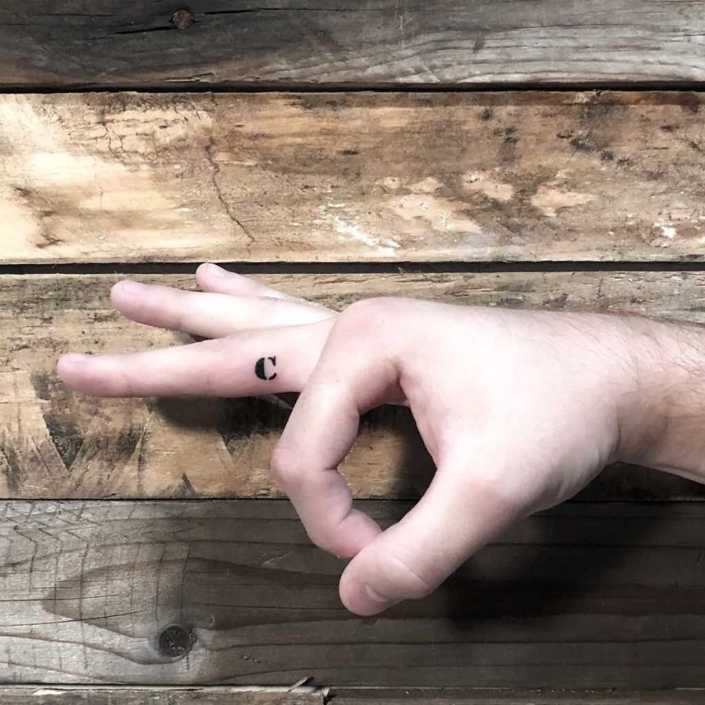 Tatuagem da letra C com letras pequenas na lateral do dedo médio