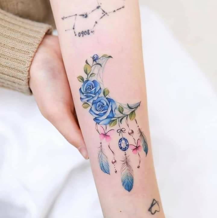 Tatuaje de rosas azules luna y llamador de angeles con gemas y mariposas en el antebrazo