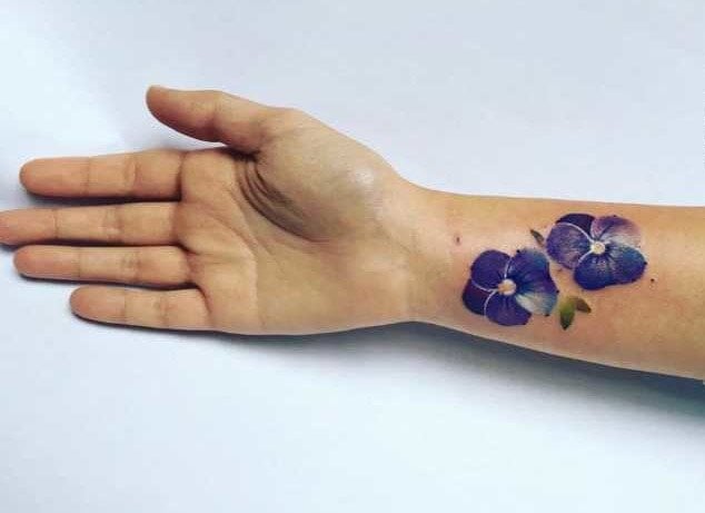 Tatuagem no pulso feminino com duas flores violetas azuis