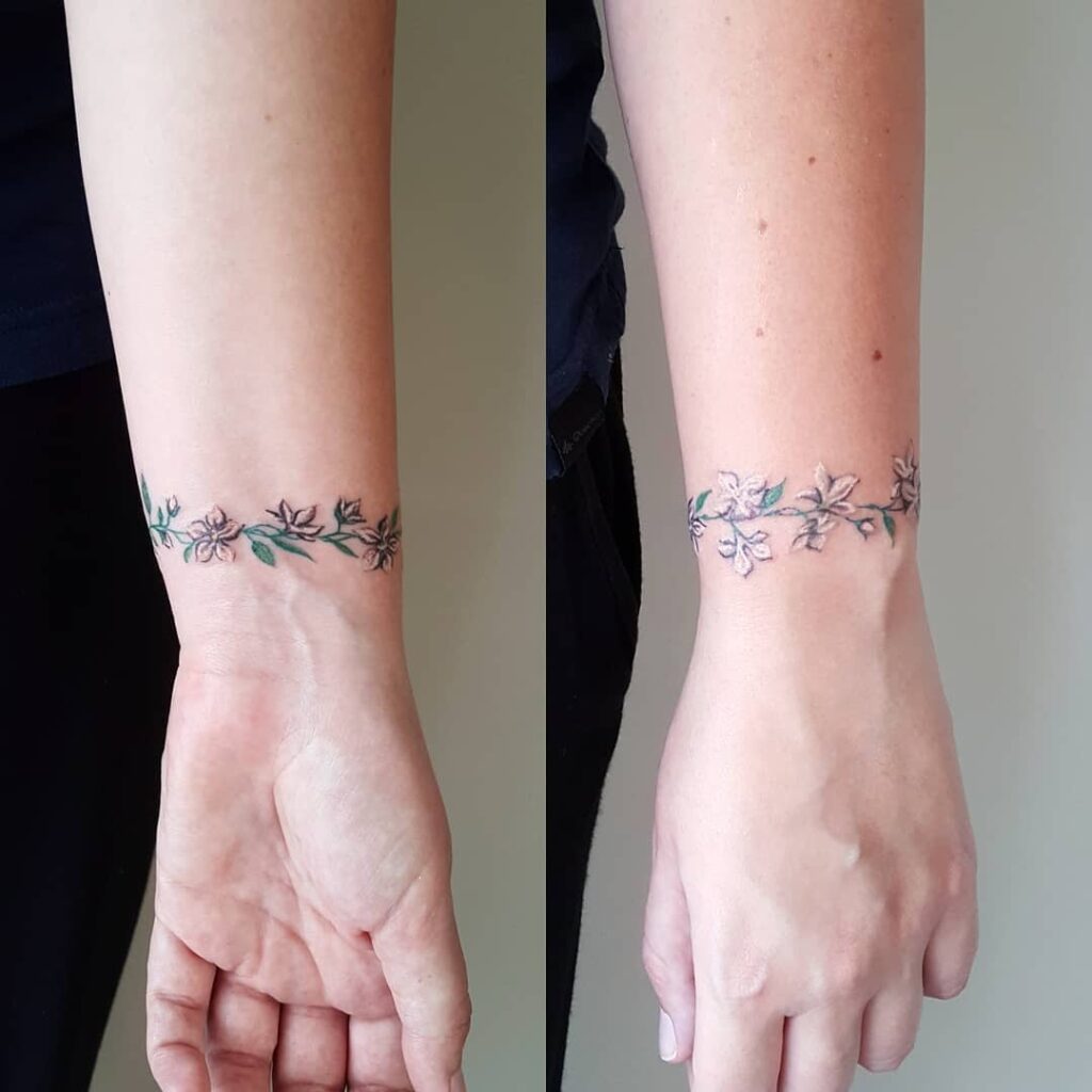 Tatuagem no pulso de um galho tipo mulher com flores brancas e rosa formando uma pulseira