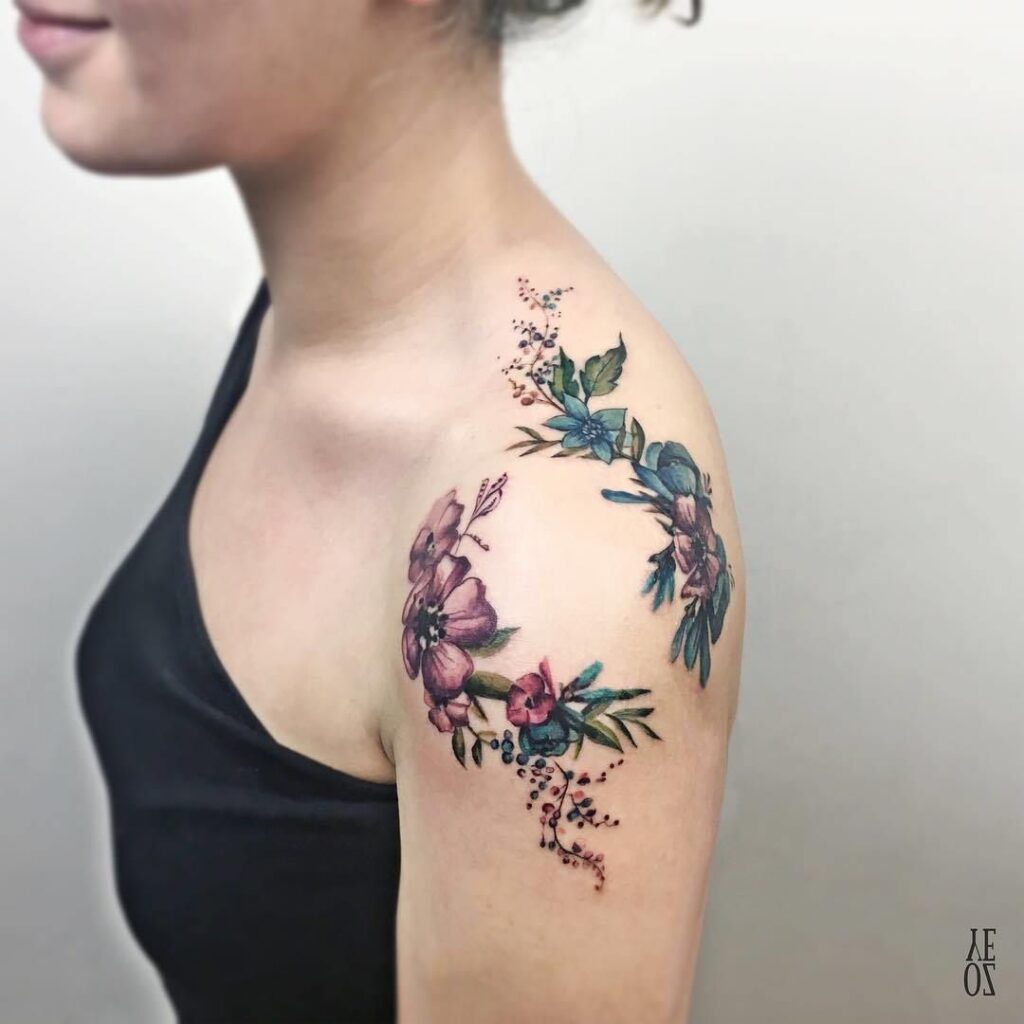 Tatuagem no ombro mulher buquês de flores violetas azuis e galhos verdes
