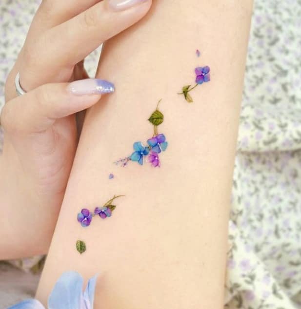 Lindas tatuagens para mulheres pequenas flores e folhas nas cores azul e violeta no braço