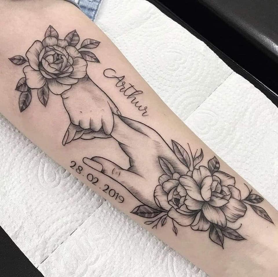 Bellissimi tatuaggi per donne: la mano del bambino che tiene la mano della madre, il nome Arthur e la data con rose nere sull'avambraccio